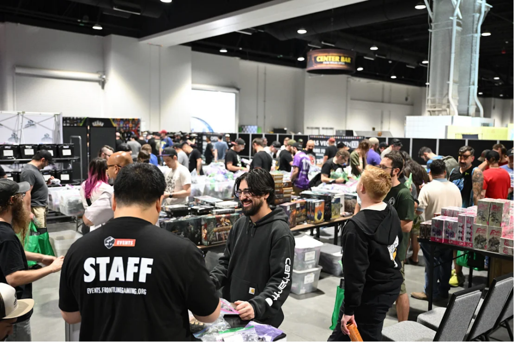 Staff member in a black STAFF shirt helps attendees at a busy convention booth with game boxes and merch on display.