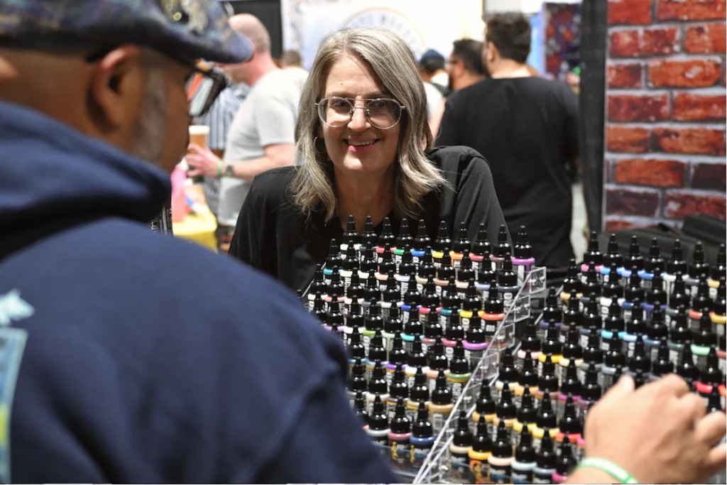 Smiling woman with glasses behind a display of colorful nail polish bottles at a market booth.