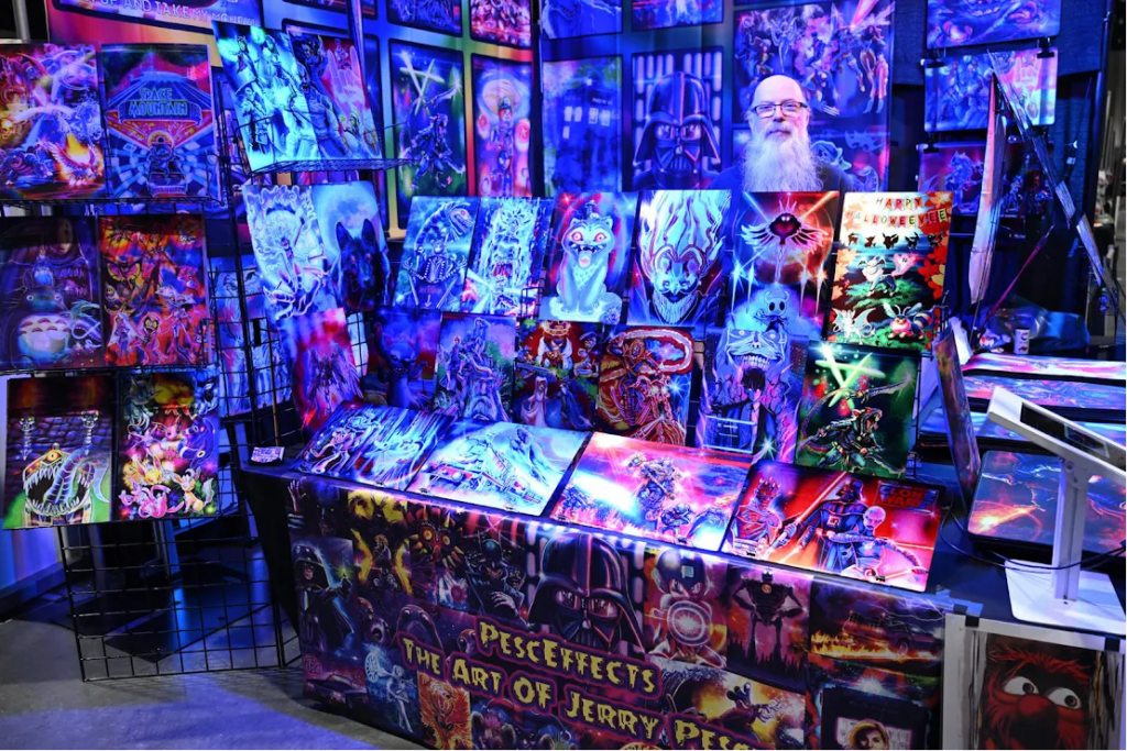 Colorful art booth with neon sci‑fi and fantasy paintings arranged on wall panels; bearded vendor behind the table at a convention display.