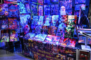 Colorful art booth with neon sci‑fi and fantasy paintings arranged on wall panels; bearded vendor behind the table at a convention display.