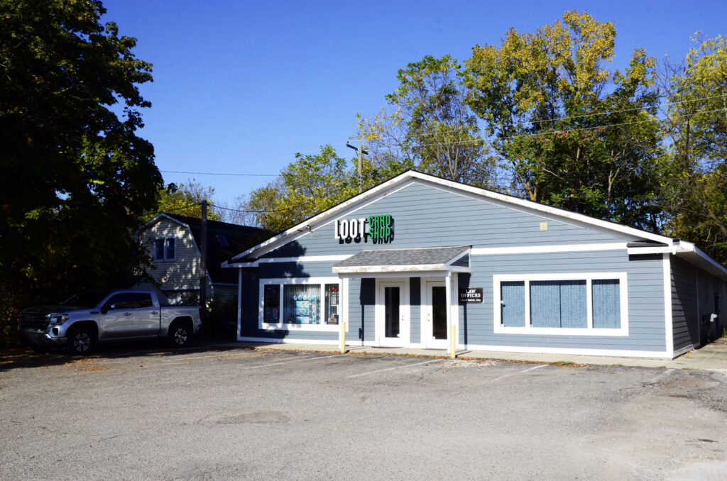 Front view of a blue storefront with a peaked roof and white trim, Loot Card Shop signage visible, in a parking lot with a silver pickup nearby.