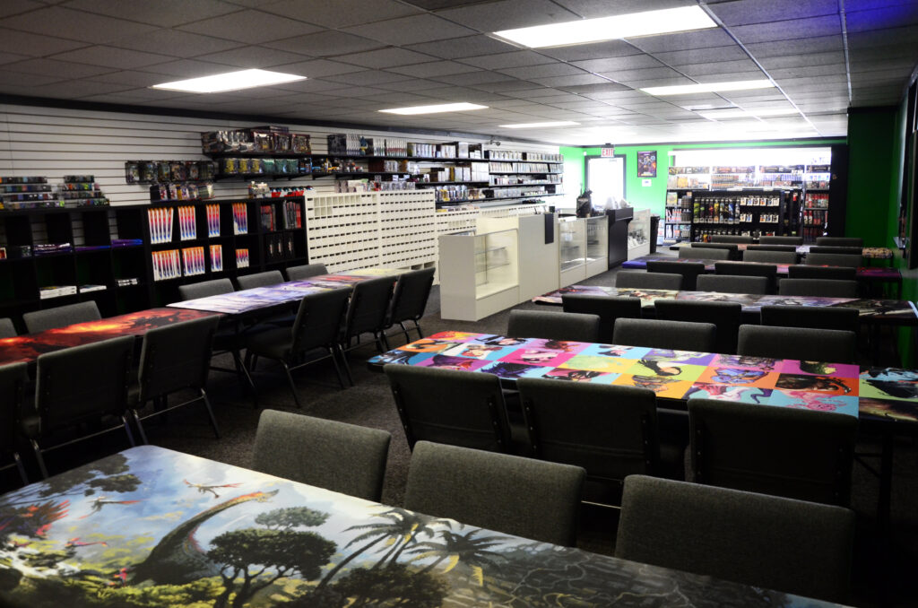 Wide view of a game store interior with long shelves of boxes along the back wall, and tables covered in colorful game mats ready for play, plus dark chairs around the tables.