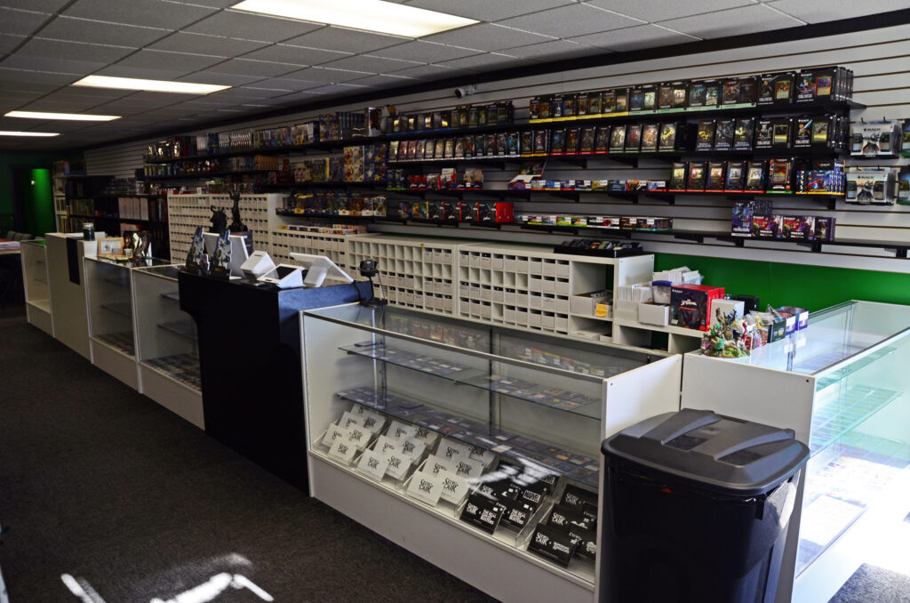 Interior of a video game store with tall shelves full of boxed games, glass display cases, a checkout counter, and a black trash bin in the foreground.