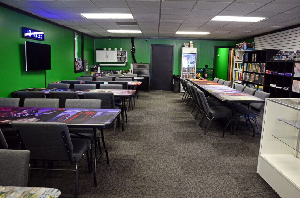 Gaming room with long tables covered in colorful mats, gray chairs, and a bright green wall. A wall-mounted TV is on the left.