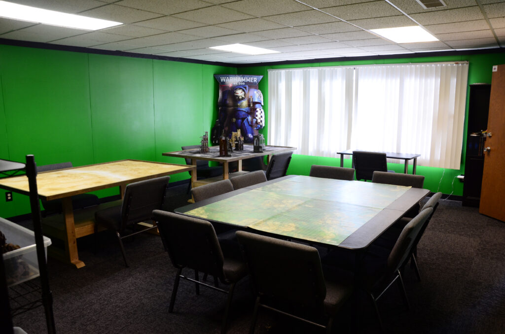Green-walled game room with long tables and miniature models on the central table, Warhammer poster on wall, and vertical blinds over a large window.