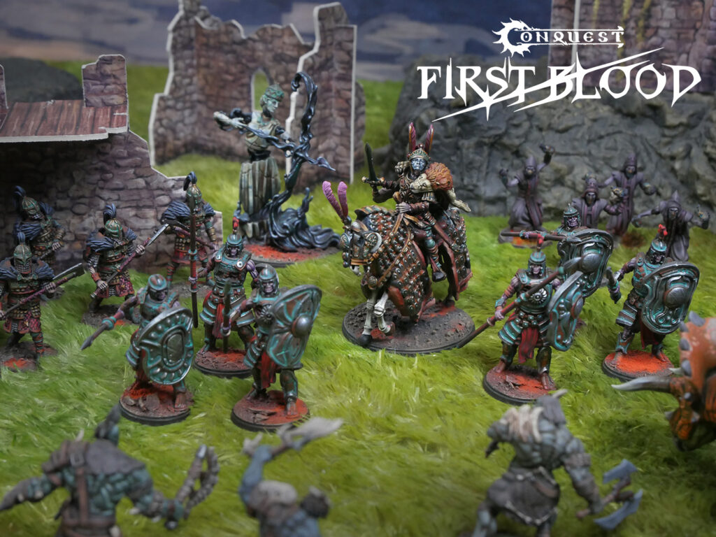 Group of painted fantasy miniatures on a grassy battlefield with ruins in the background and a mounted hero in ornate armor near center-right, logo 'Conquest First Blood' visible in the sky area.5 characters]