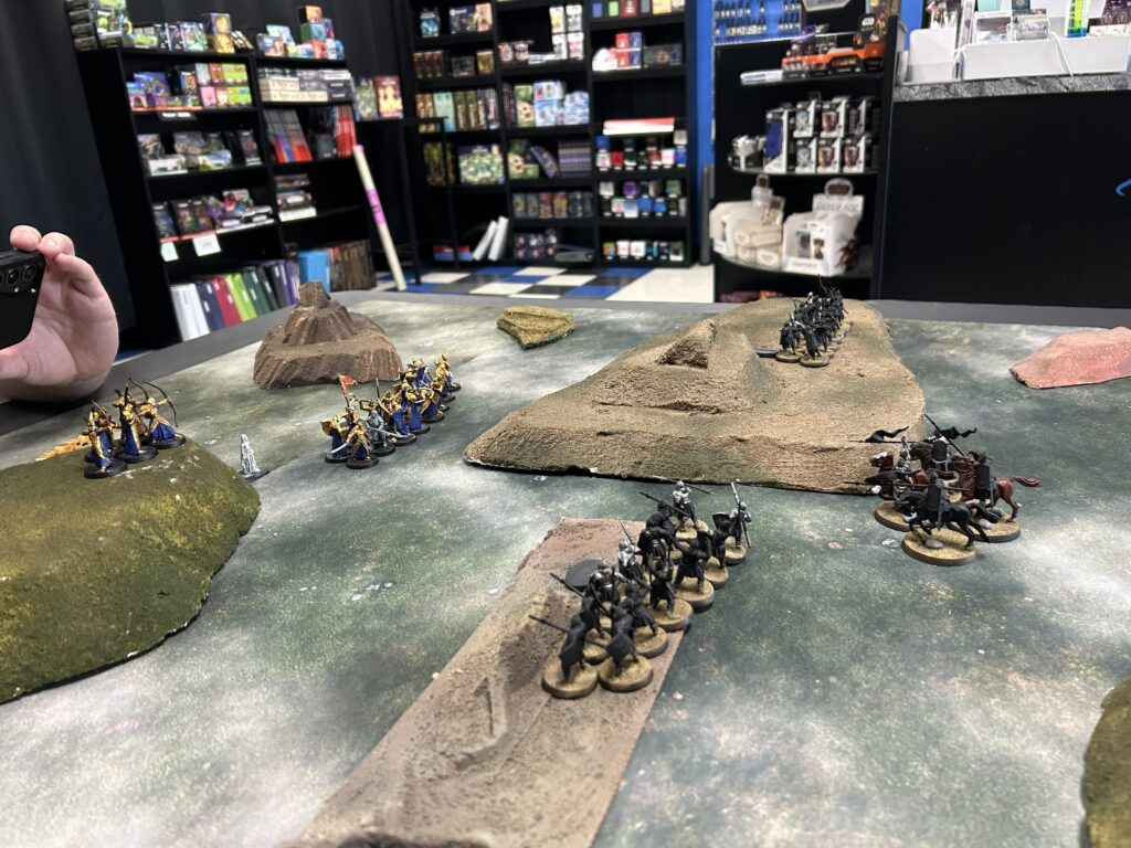 Tabletop wargaming scene with multiple groups of miniature soldiers on rocky terrain, a person holds a phone on the left, and shop shelves in the background.
