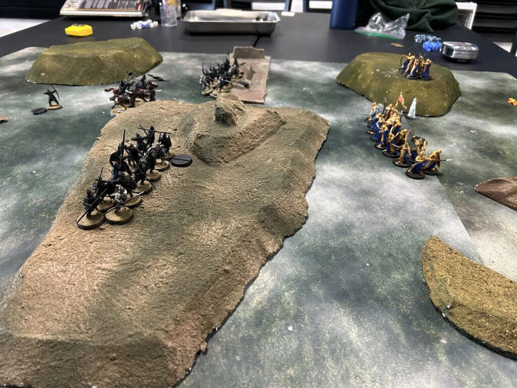 Tabletop wargame scene with two opposing armies lined up on a rocky, hilly battlefield.