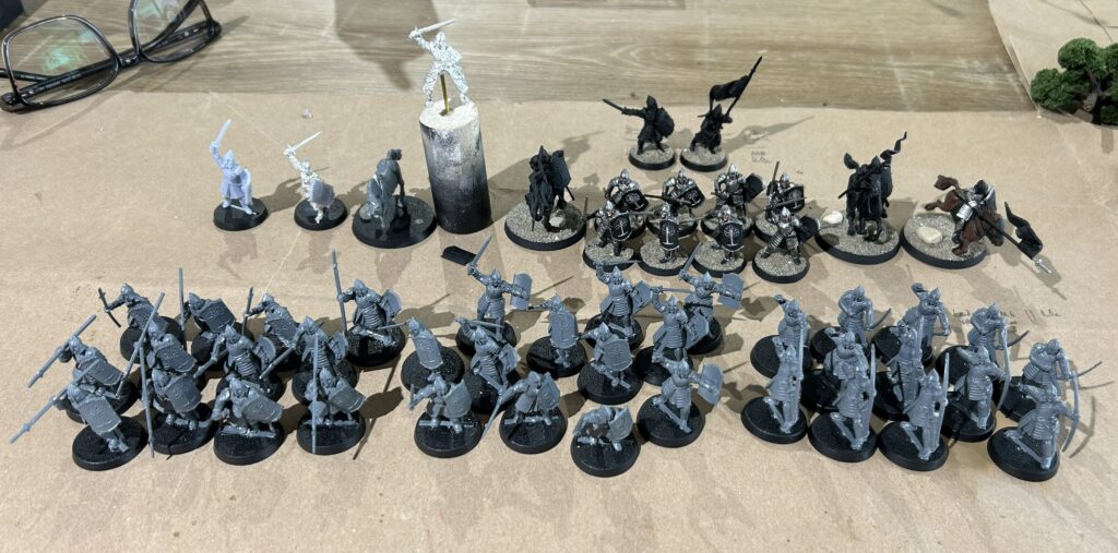 Row of gray unpainted fantasy miniatures in front, with darker metal figures to the rear, all set on a brown work surface with glasses nearby.