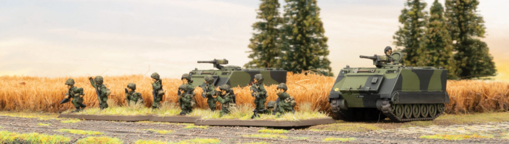 image Frontline Gaming Row of miniature soldiers advancing beside camouflaged armored vehicles on a model battlefield in a grassy field.