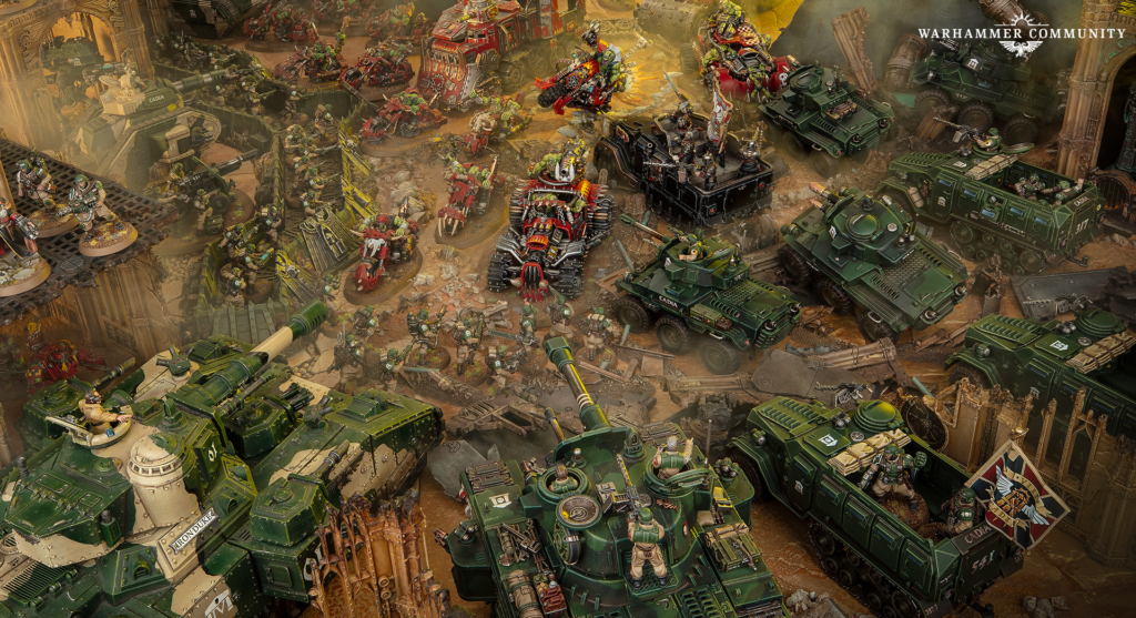 Wide Warhammer 40k battle scene with green armored tanks, walkers, and infantry clashing amid a ruined battlefield.