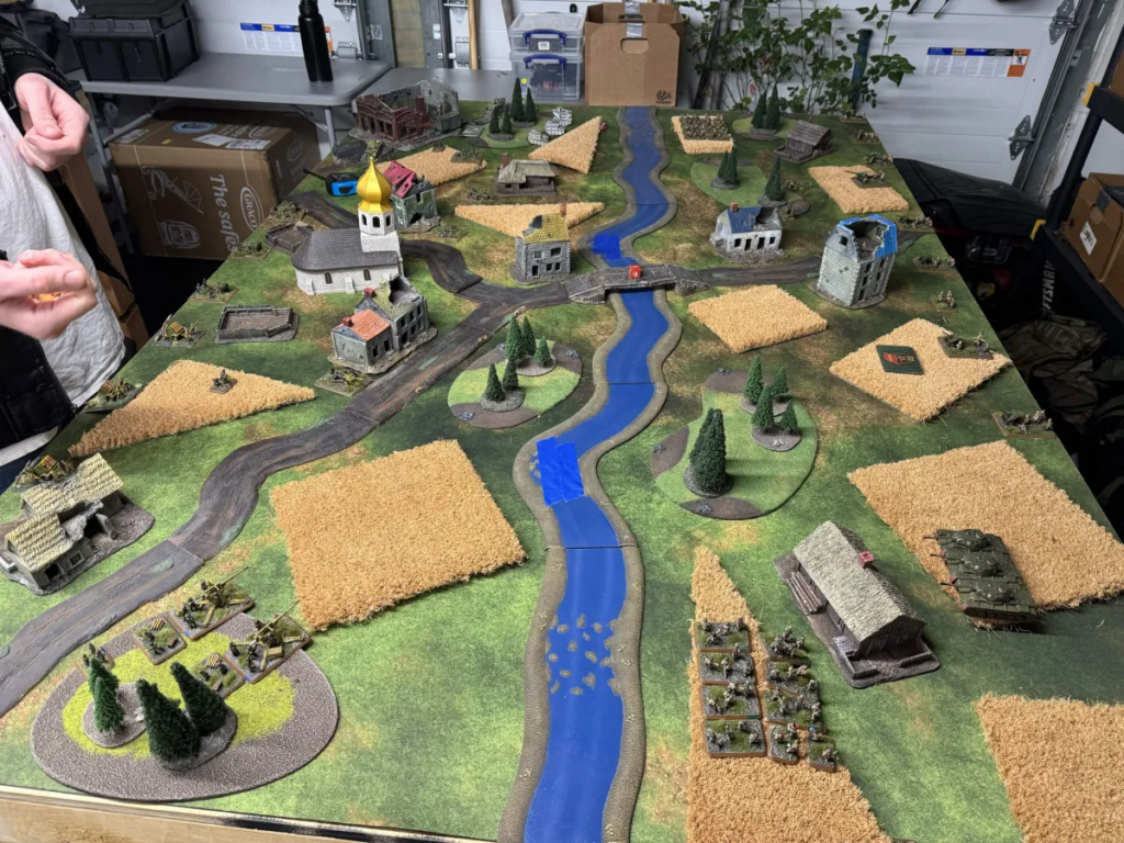Tabletop wargame battlefield with a winding blue river, roads, fields, trees, and miniature houses and soldiers.