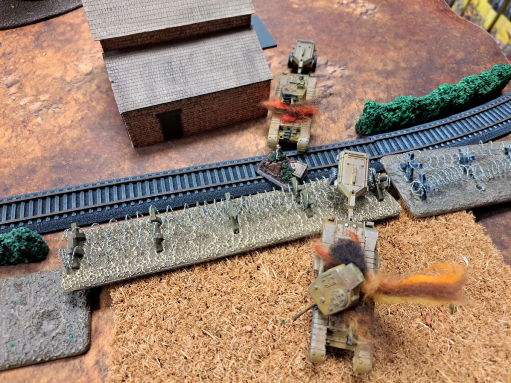 Tabletop wargaming scene with tan armored vehicles advancing past a brick building, barbed wire, and a curved railway track on brown terrain.