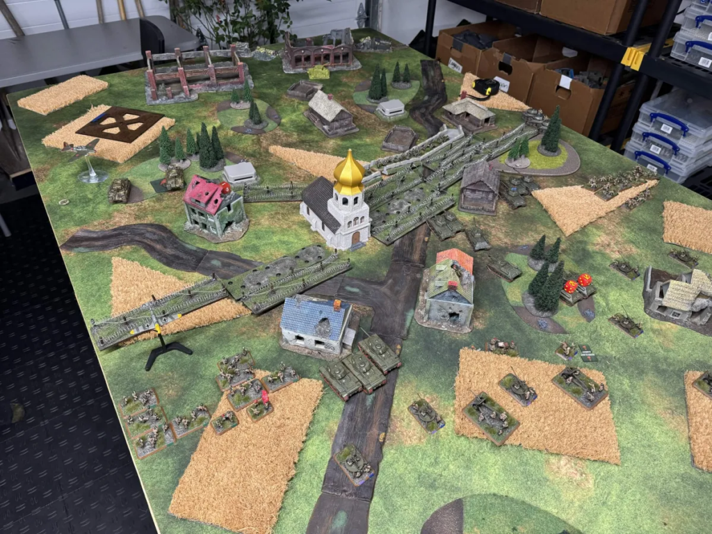 Tabletop wargame battlefield with miniature tanks, infantry, and a central church on green terrain with roads and fields
