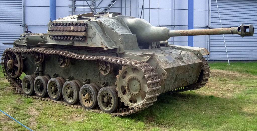Armored tank with a long gun barrel, muddy tracks, and multiple road wheels on grass beside a blue building.