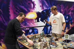 Two men play a tabletop wargame at a cluttered battle table, with miniatures and ruined buildings, in front of a vibrant space-themed mural.