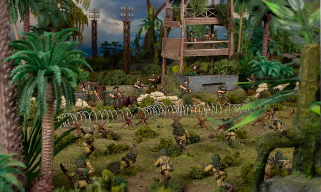 Miniature jungle battlefield with soldiers advancing behind barbed wire and a wooden watchtower.