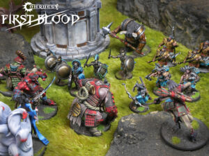 Tabletop fantasy battle with armored miniatures clashing on a grassy battlefield beside a stone ruin; Conquest First Blood logo visible in the corner.