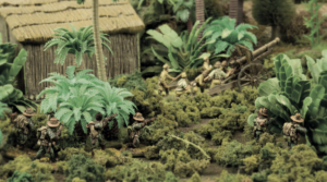 Miniature war diorama: soldiers advancing through jungle toward a cannon near a thatched hut.
