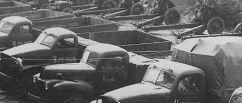 Black-and-white photo of a row of vintage 1940s pickup trucks parked in a scrapyard or yard.