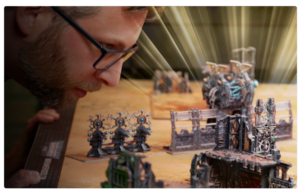 A man wearing glasses leans close to a tabletop wargaming battlefield with painted miniatures and ruined structures.