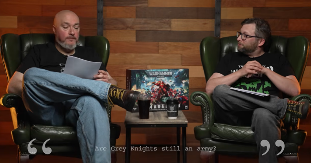 Two men in green leather chairs discuss on a studio set; a Warhammer 40K box sits on the table between them, caption: 'Are Grey Knights still an army?'