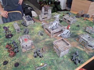 Tabletop wargame battle scene with futuristic miniatures on a green terrain mat and modular ruined buildings, dice scattered around.