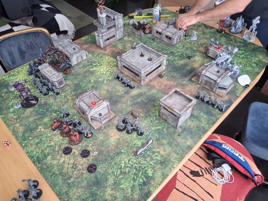 Tabletop wargame battle scene with gray industrial buildings, urban ruins, and infantry miniatures on a grassy mat.