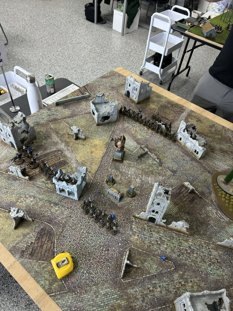 Tabletop wargame setup with ruined stone buildings, armies lined up for battle on a cobblestone map, and measuring tape nearby.