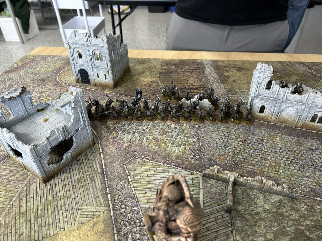 Tabletop wargame scene with ruined stone castle walls and a line of medieval miniature figures on a cobblestone battlefield.