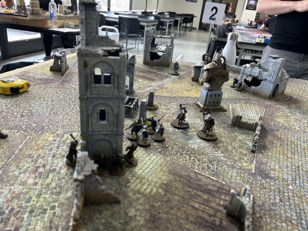 Tabletop wargame battle scene with medieval ruins and miniature soldiers on a textured game board.