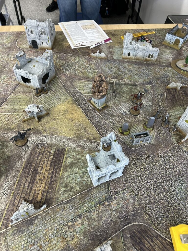 Tabletop wargame scene with ruined stone buildings, cobblestone streets, a central statue on a pedestal, and scattered miniature figures; a rulebook and dice on the board.