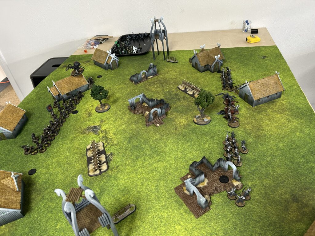 Tabletop wargame setup with huts, ruins, trees, and two armies of miniature figures on a green gaming mat behind the battlefield.