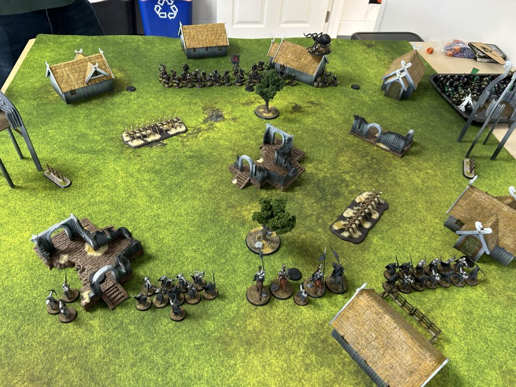 Tabletop wargame scene on a grassy mat with rows of armored miniatures, huts, and ruined buildings arranged for battle.