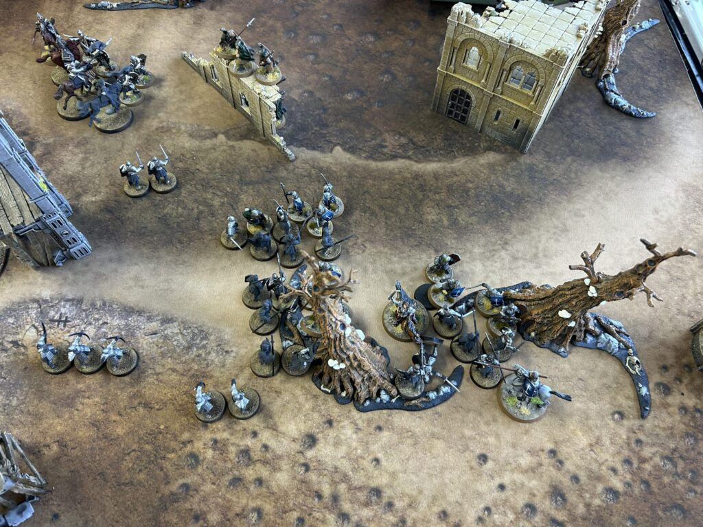 Tabletop fantasy battle scene with groups of armored miniatures near ruined stone walls and a large ruined building on a textured gaming mat.