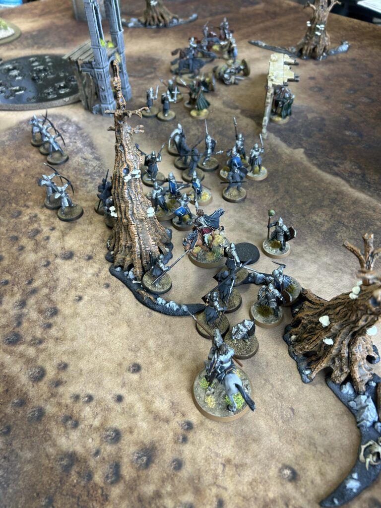 Tabletop wargame scene with gray-armored miniatures gathered around a twisted tree and ruined structures on a textured battle mat.