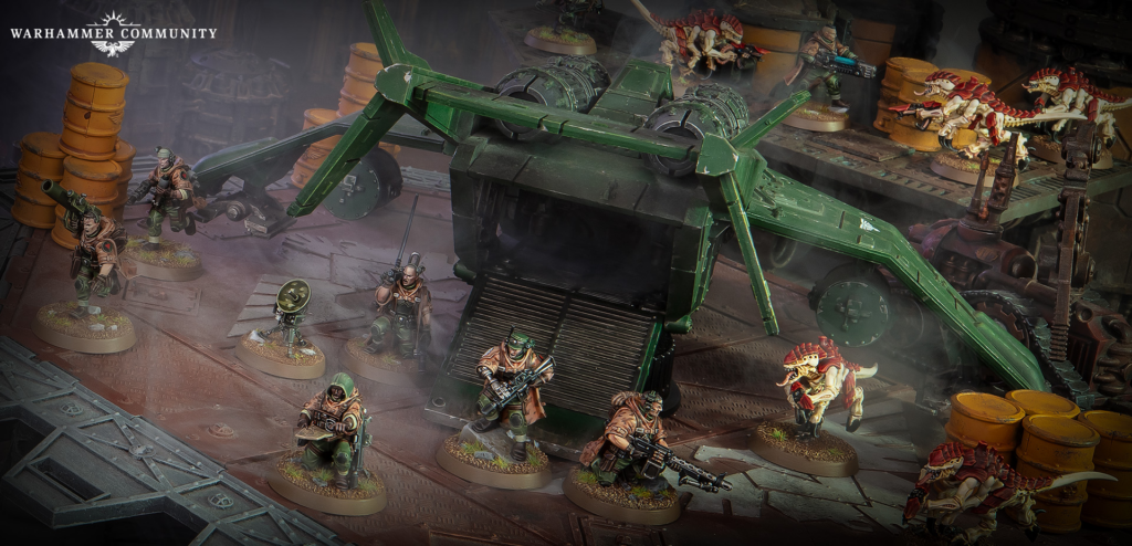 Warhammer Community banner over a tabletop battle scene with a large green aircraft centerpiece and miniatures engaging reptilian enemies in a ruined industrial setting.