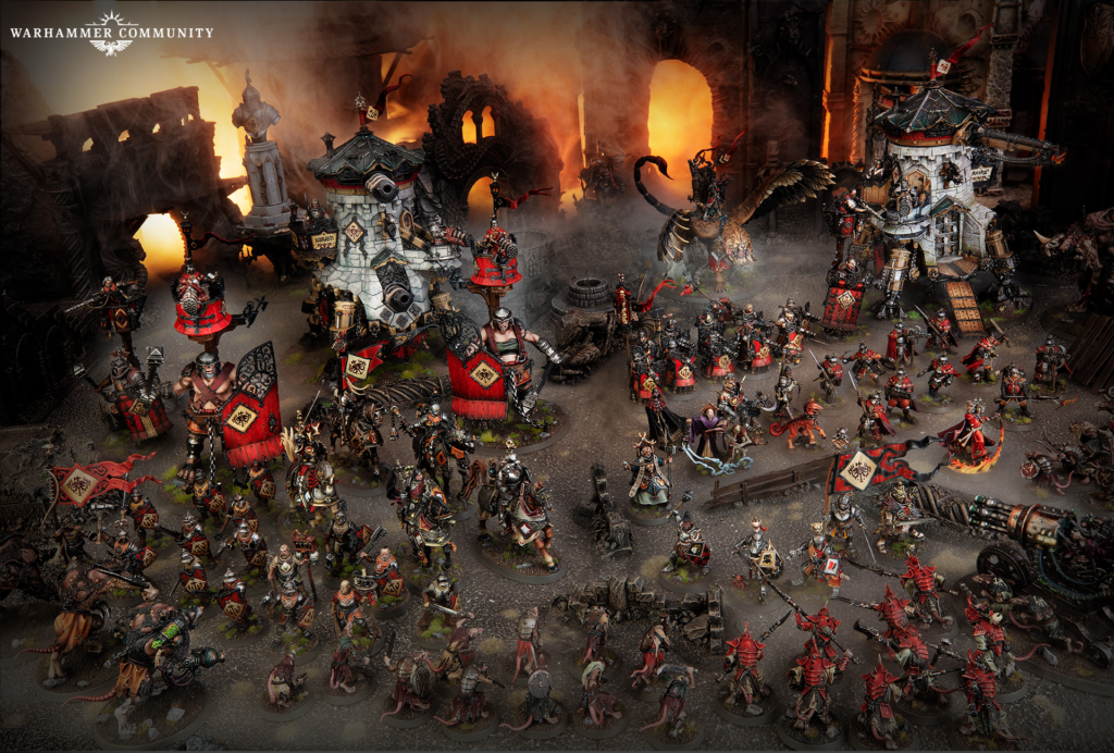 Large Warhammer battle diorama with numerous red-armored soldiers, banners, and siege engines amid a fiery ruined fortress.