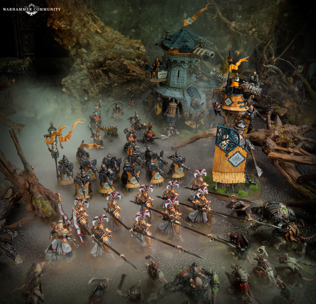 Warhammer Community logo in top-left with a massive tabletop battle scene of armored troops marching through mist around a siege engine fortress.