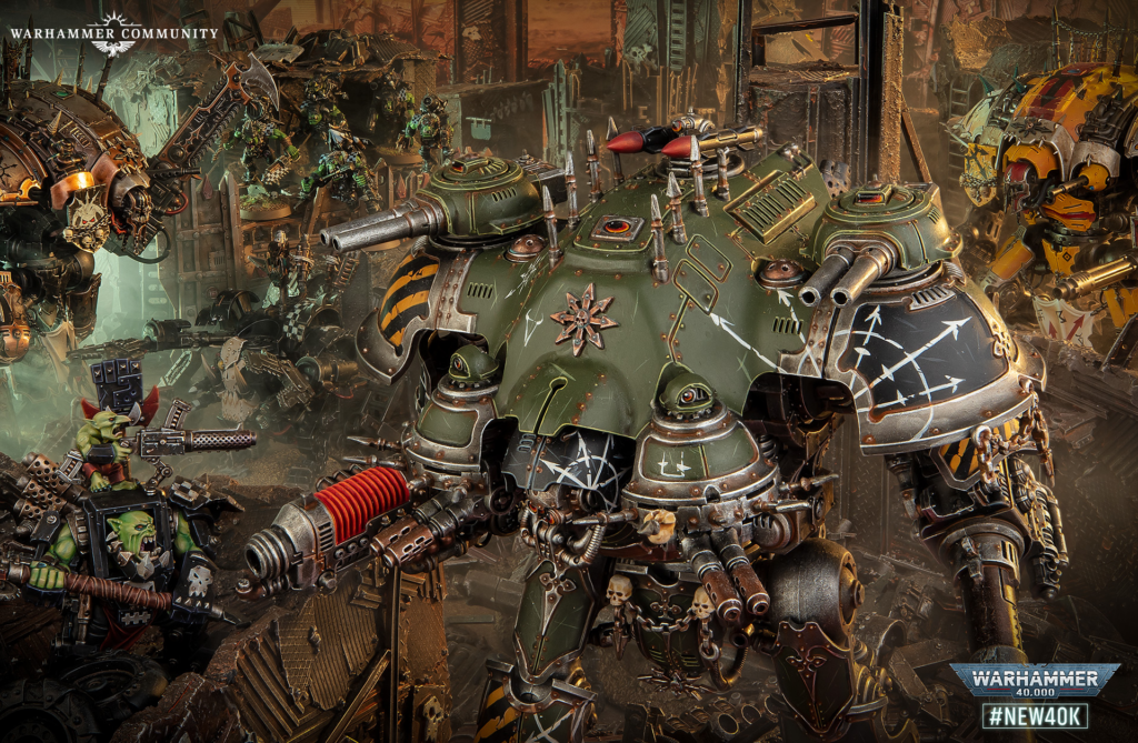 A giant green Warhammer 40k siege walker leads orc forces through a ruined city, with weapons firing and debris everywhere, Warhammer Community logo bottom-right.