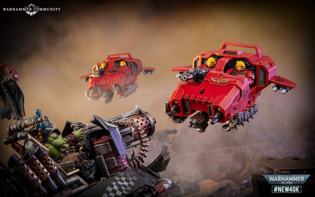 Red armored battle tank with yellow pilots fires dual cannons in a smoky battlefield, looming wreckage in foreground, Warhammer branding visible.
