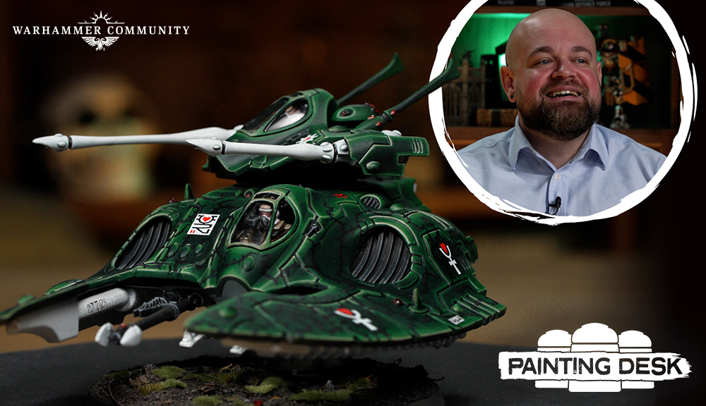 image Frontline Gaming Green Warhammer tank model on a table, Warhammer Community logo in top-left; inset circle shows a smiling man in a blue shirt; Painting Desk logo bottom-right.
