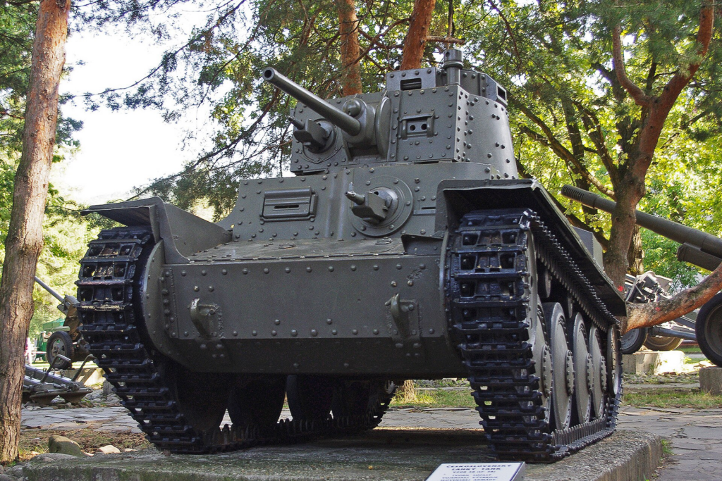 image Frontline Gaming World War II-era tank on display outdoors in a park among trees, with a long gun barrel and riveted armor.