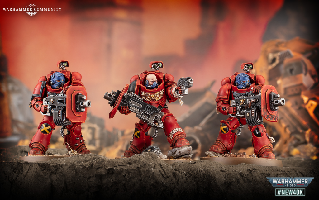 Three red-armored Space Marines pose with large weapons on a rocky battlefield diorama; fiery, orange background and distant debris.A watermark in the bottom-right reads 'Warhammer 40,000 #NEW40K'.
