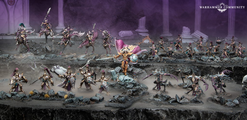 Large diorama of Warhammer fantasy miniatures clashing on a ruined rocky battlefield with purple fog and ancient columns in the background. Central golden-armored hero with purple cape leads the charge with a sword and shield amid other ornate figures.