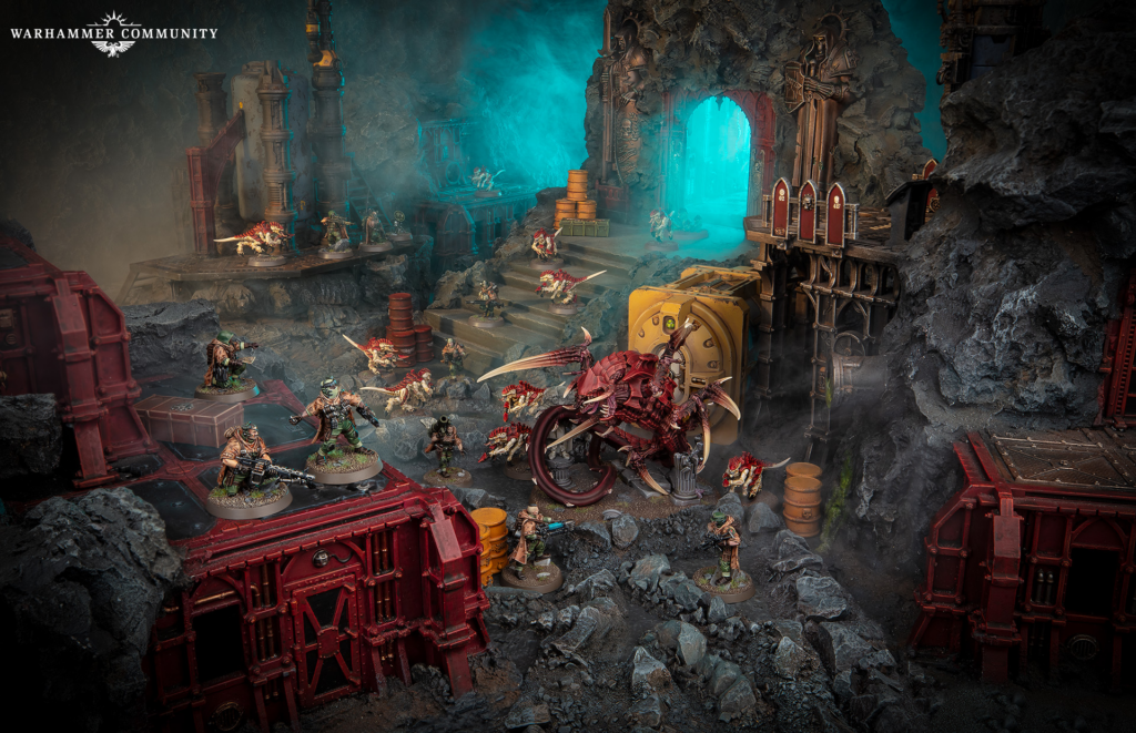 Tabletop Warhammer battle diorama in a rocky cavern with red modular ruins, teal glowing portal, and many painted miniatures engaged in combat nearby.