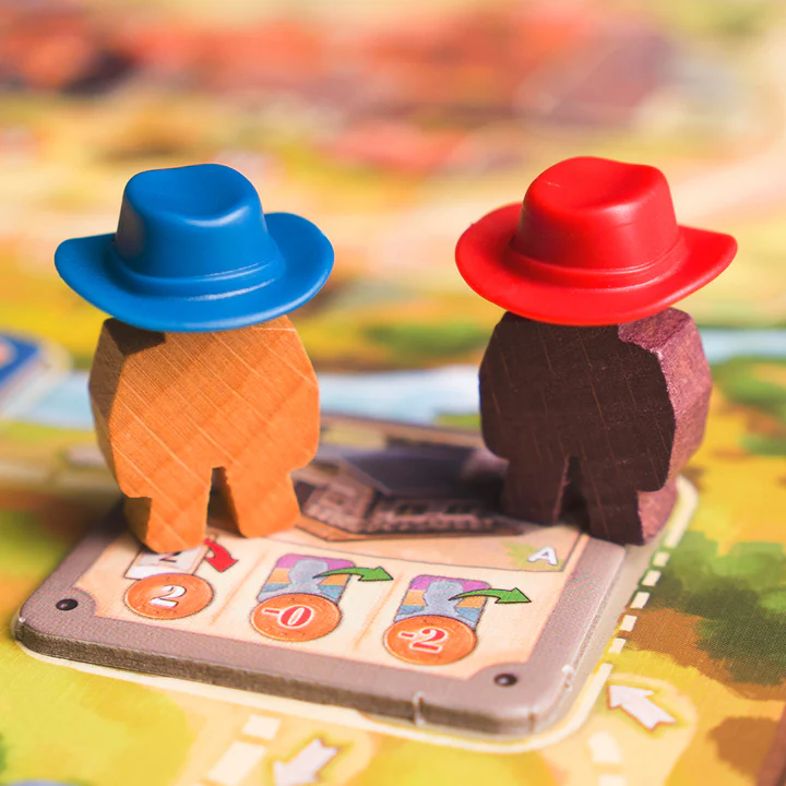 image Frontline Gaming Two wooden game pawns with blue and red hats stand on a colorful board game tile with action cards and tokens visible beneath them.