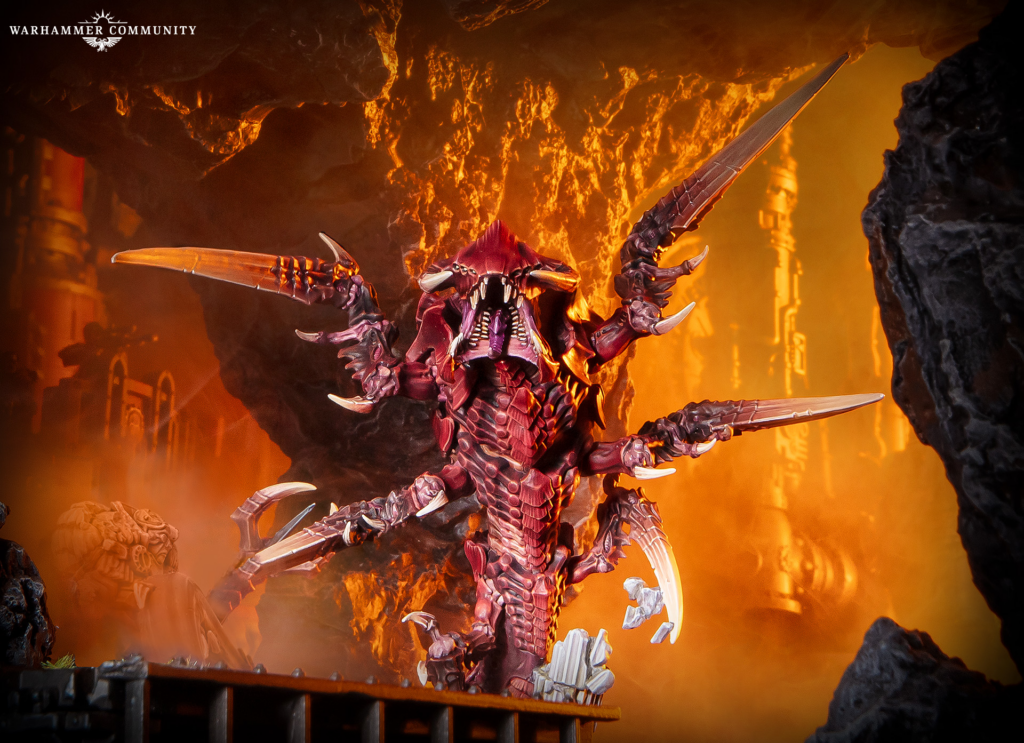 Red-painted dragon-like model with large wings and jagged armor stands in a fiery lava cave setting for Warhammer, glowing orange surroundings.