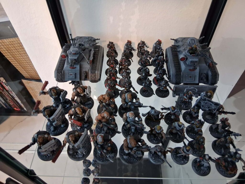 Row of grey armored tanks flanked by groups of sci-fi infantry miniatures on a glass display shelf.","Warhammer-style miniature army: two tanks at ends and a line of infantry figures in the middle.","Collection of grey-painted battle robots and soldiers arranged on a glass shelf for display.","Grey armored tanks with infantry miniatures on display case, ready for tabletop battles.","Display of miniature wargaming figures: two large tanks on either side and soldiers in the center."]} }{