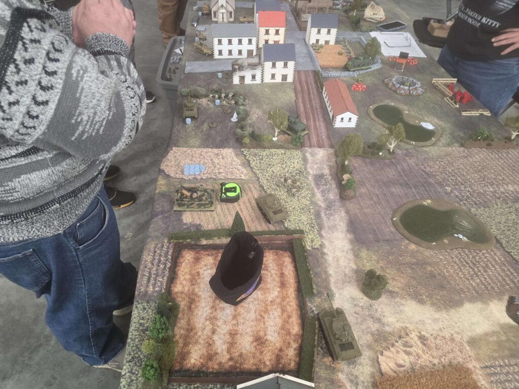 Top-down view of a tabletop wargame with miniature buildings, tanks, and hills on a textured mat.
